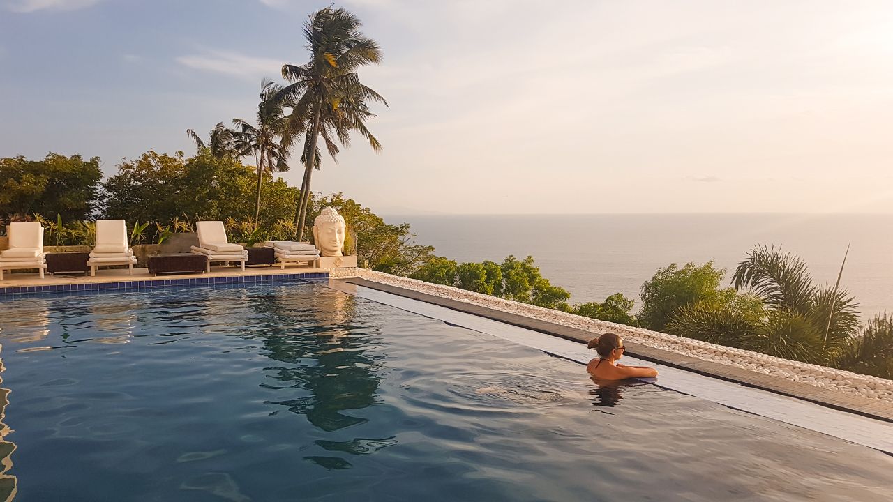 A young woman leaning at the edge of an infinity pool and looking at the sun setting into the sea. There is a big palm tree in front. Calmness and serenity. Luxury hotel in Lombok, Indonesia.