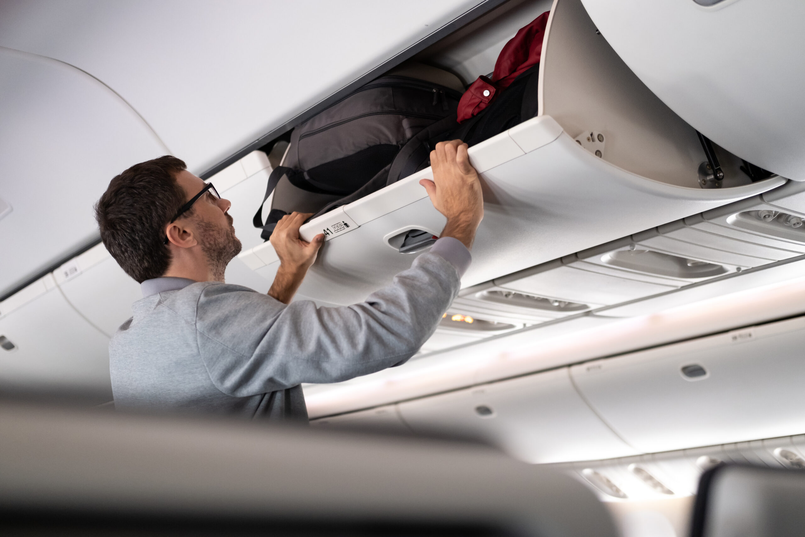 Young man putting luggage into overhead locker on airplane. Traveler placing carry on bag in overhead compartment in aircraft