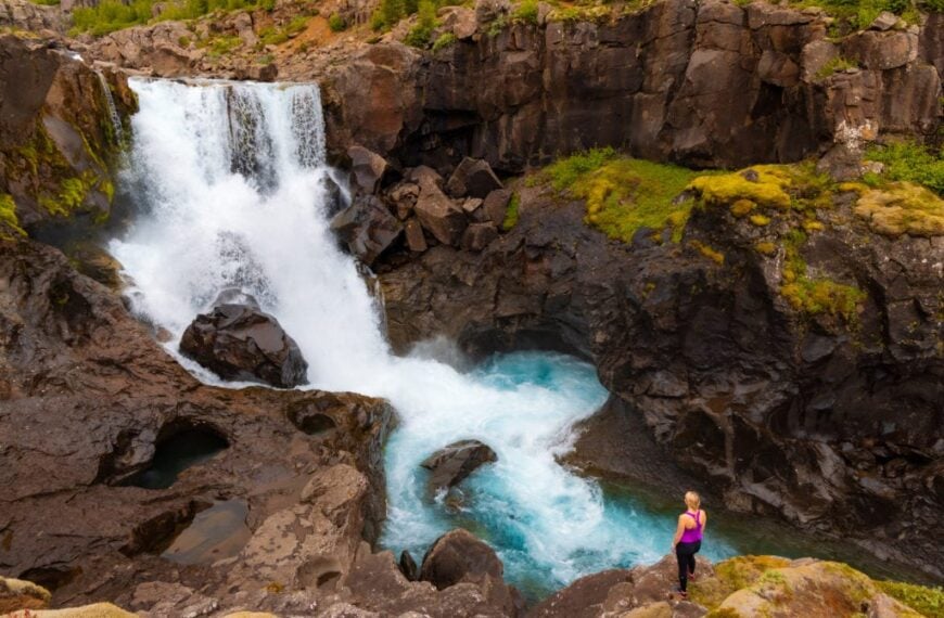 A hiker stands on the edge with a waterfall in front of her.