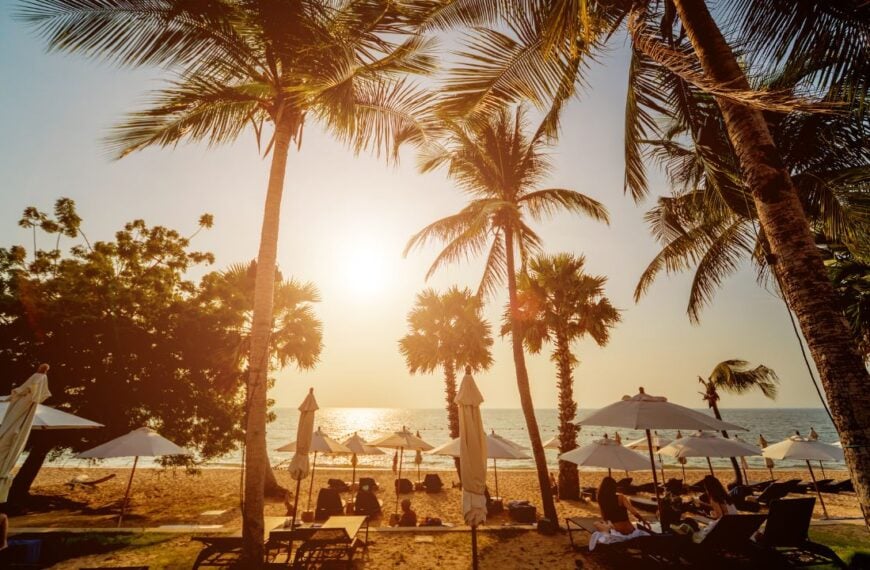 Palm trees frame a golden sunset over a white-sand beach lined with lounge chairs, capturing the relaxed, upscale atmosphere of Florida’s Gulf Coast resort living