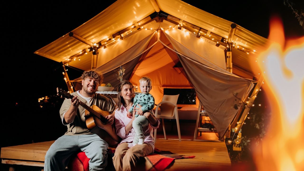 Happy family relaxing and spend time together in glamping on summer evening and playing guitar near cozy bonfire. Luxury camping tent for outdoor recreation and recreation. Lifestyle concept