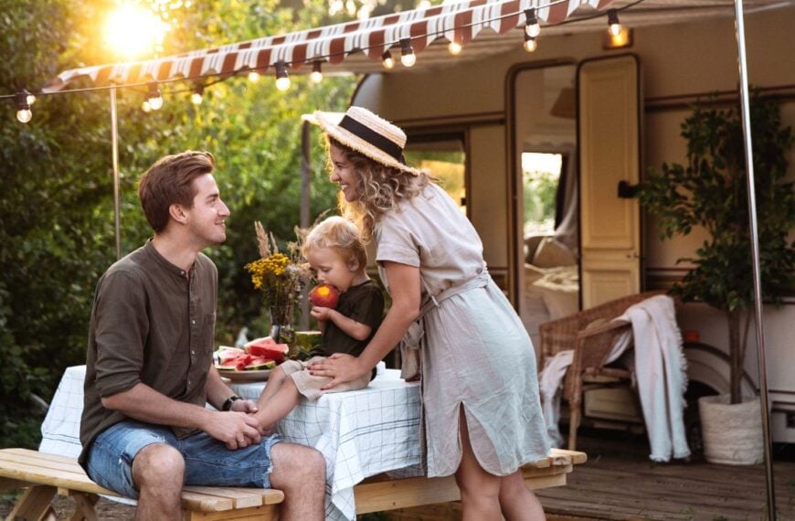 Family vacation in mobile home: young parents travel with small preschool son have picnic on terrace on sunset at rv camper trailer. People enjoy summer road voyage on caravan car. Summer outdoor trip