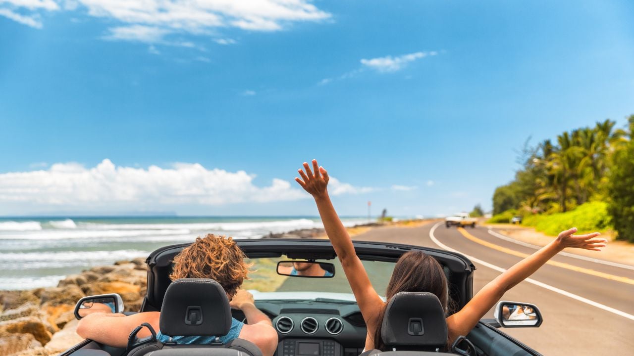 Road trip car holiday happy couple driving convertible car on summer travel Hawaii vacation. Woman with arms up having fun, young man driver