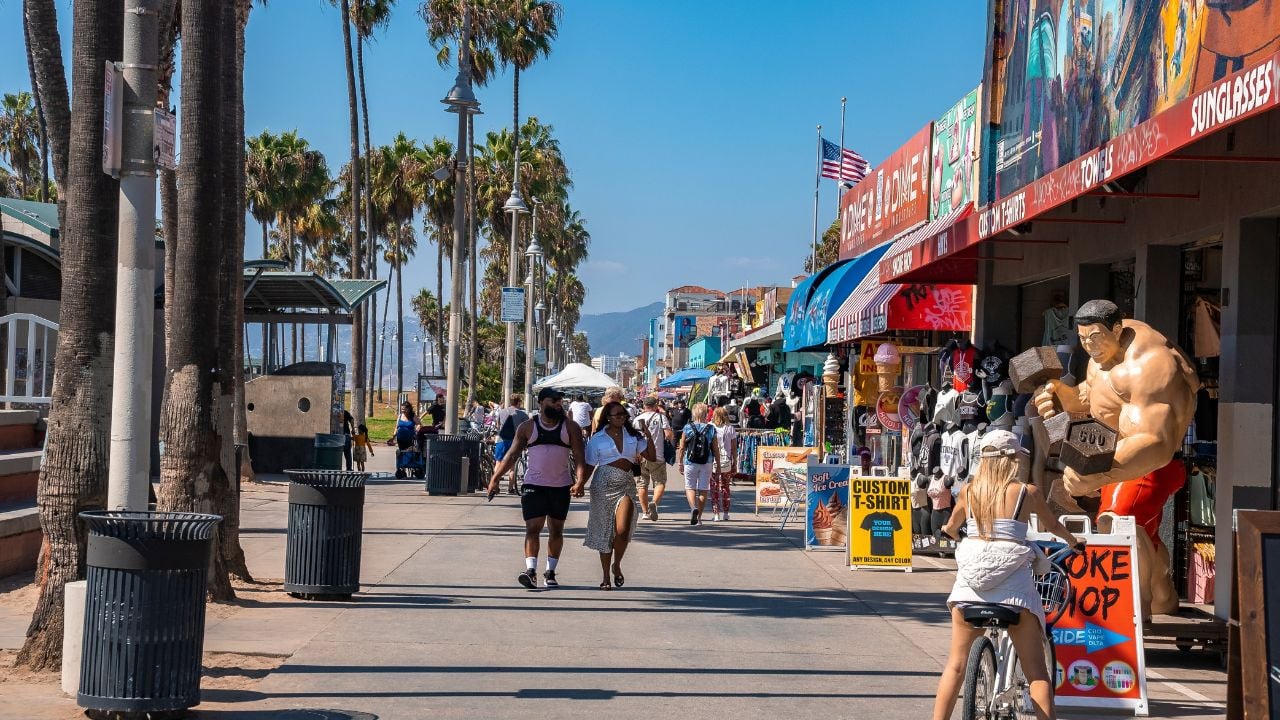 Los Angeles, USA. September 20, 2022. Tourists walking on boardwalk by palm trees and exploring shops in a row at Venice beach with clear blue sky in background during sunny day