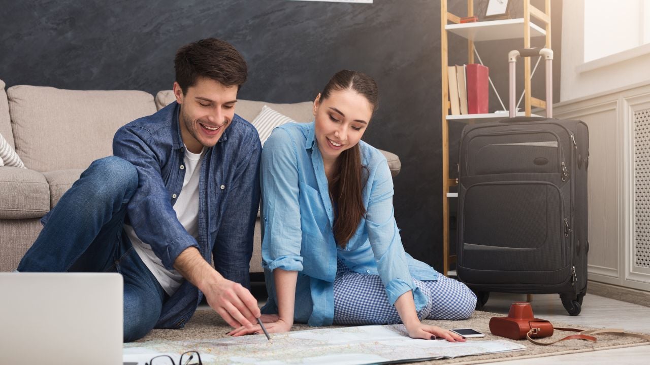 Couple planning vacation trip, studying map, sitting on floor at home with laptop, copy space