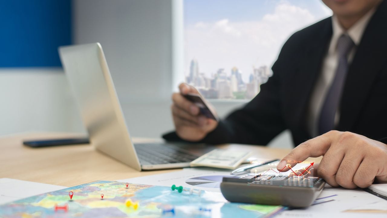 Close-up of a businessman using a calculator, laptop, and credit card while planning travel expenses with a world map on desk.