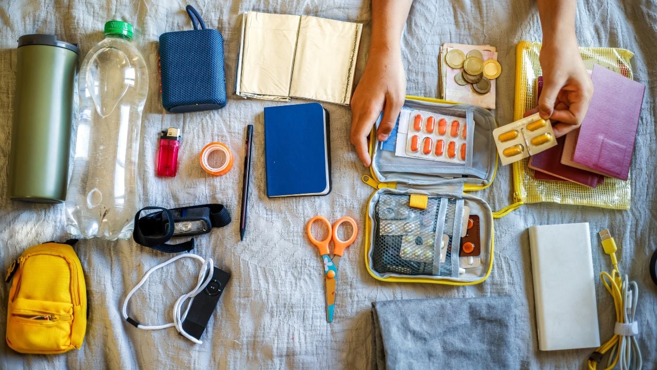 Blister with pills in hands of white middle-aged woman, assembling and checking emergency go-bag on blanket in bedroom at home close-up: scissors, thermal blanket, pills, first aid kit, power bank