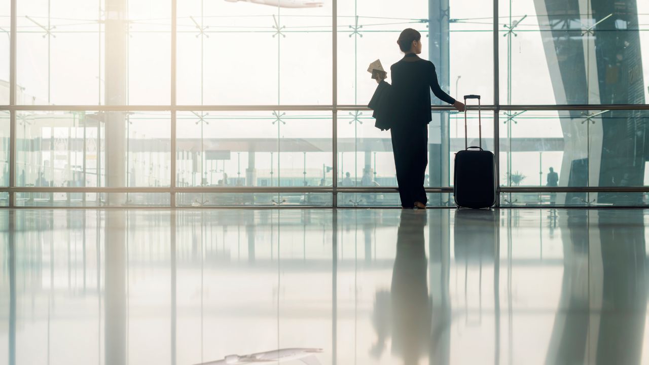Young elegant business woman in international airport looking out window at flying airplane while waiting at boarding gate before departure, holding passport and boarding passes in her hand.