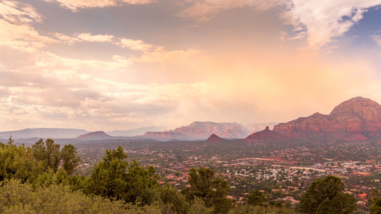 A wide scenic view over Sedona, Arizona, with red rock formations glowing in warm early light beneath a soft, cloud-filled sky