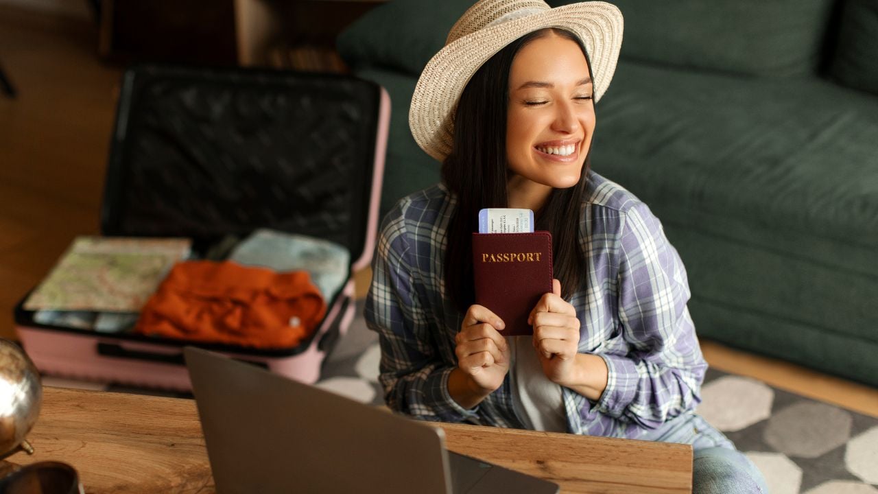 Excited happy woman holding passport and tickets, sitting in front of laptop with open suitcase on background, preparing for abroad tourist trip