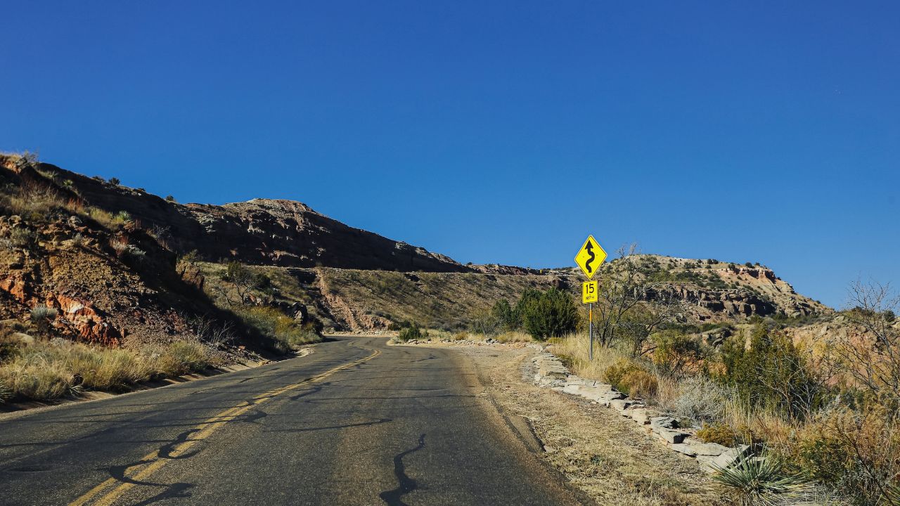 Winding road in Palo Duro Canyon. Texas desert landscape. Curved asphalt through rocky hills. Blue sky background. Scenic travel route. American southwest nature. Empty road journey scene.