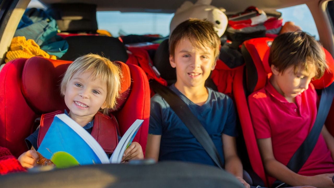 Children traveling in car seats, reading and using a device.