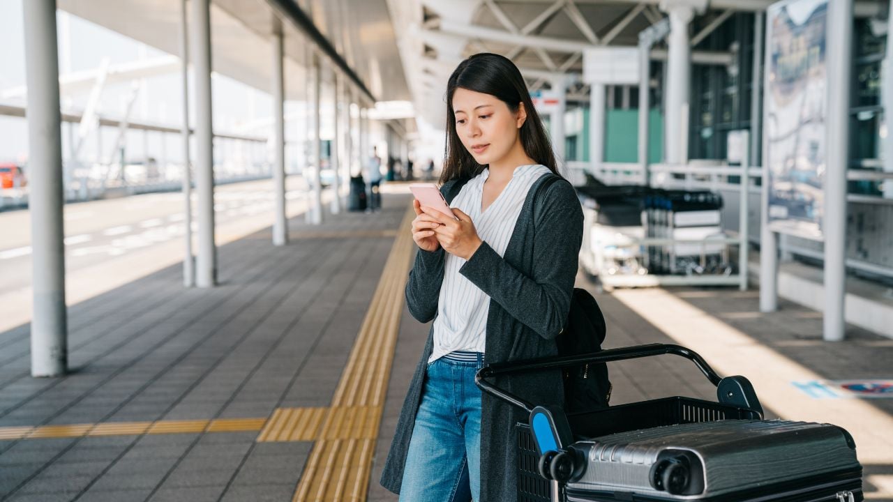 Young Asian passenger standing beside luggage cart and using a cellphone at the airport.