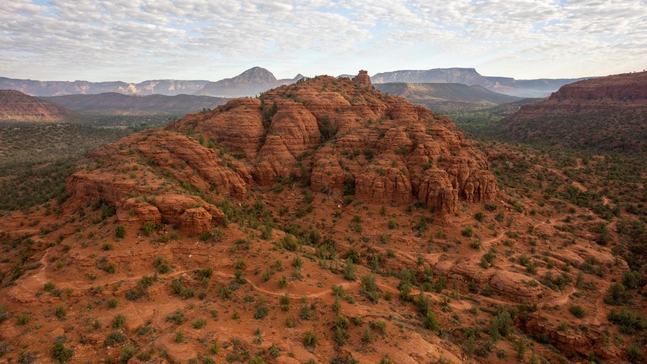 A broad aerial view of Sedona’s red rock terrain, featuring layered sandstone hills, desert vegetation, and distant mesas beneath a textured cloud-filled sky. This expansive Arizona desert landscape emphasizes scale, natural patterns, and rugged beauty, making it ideal for travel, nature, environmental, and outdoor lifestyle visuals.