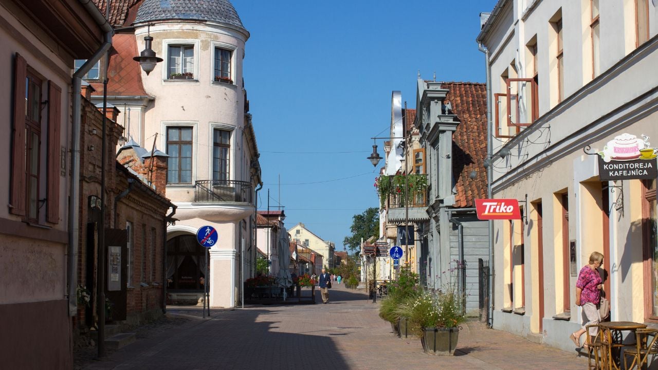 Central street in Kuldīga, Latvia, with pastel buildings, pedestrians, and a church dome in the distance