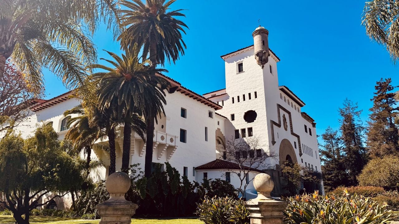 The Santa Barbara County Courthouse stands majestically with its white stucco walls, red-tiled roof, and tower framed by lush palm trees and gardens under a clear blue sky.
