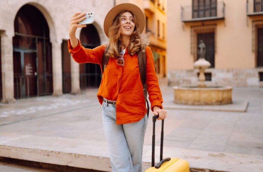 Cheerful female tourist in hat and carrying yellow suitcase strolls along city streets with phone. Young woman with phone is blogging, enjoying trip on sunny day. Concepts for blogging, weekends.