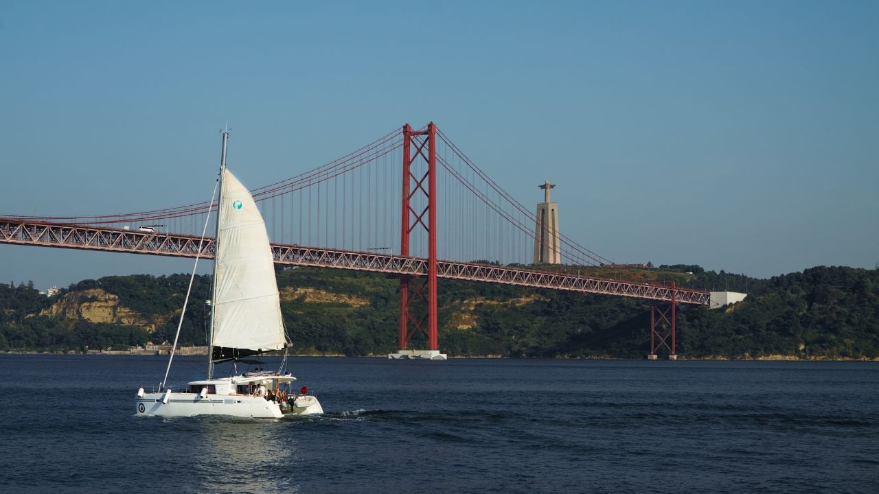 LISBON, PORTUGAL - JUNE 16 2025:Sailing catamaran sails under 25 April Bridge against of statue of Christ on opposite bank. Concept of summer, relax, cultural heritage, architecture, water transport