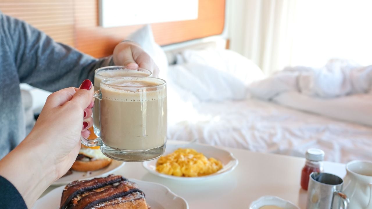 Two people toasting with coffee mugs over a breakfast table in a hotel room.