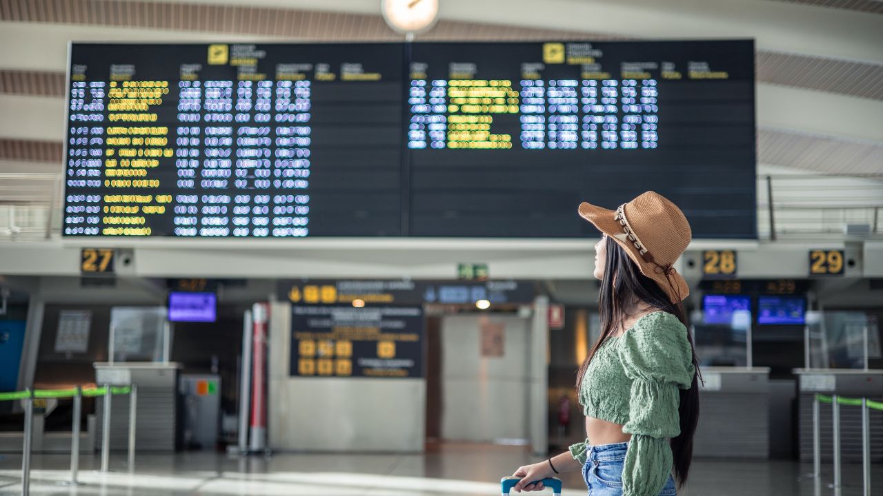 Hispanic girl looking at flight screens in airport terminal while carrying her luggage copy space