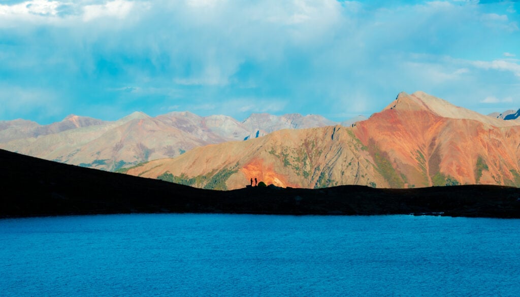 two people stand on a ridgeline across a lake with mountains behind them
