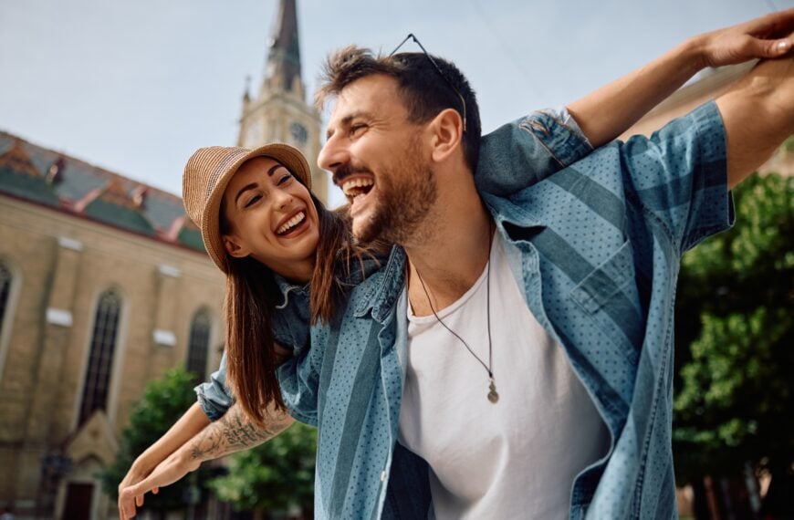 Playful couple of travelers having fun while piggybacking with arms outstretched in the city.