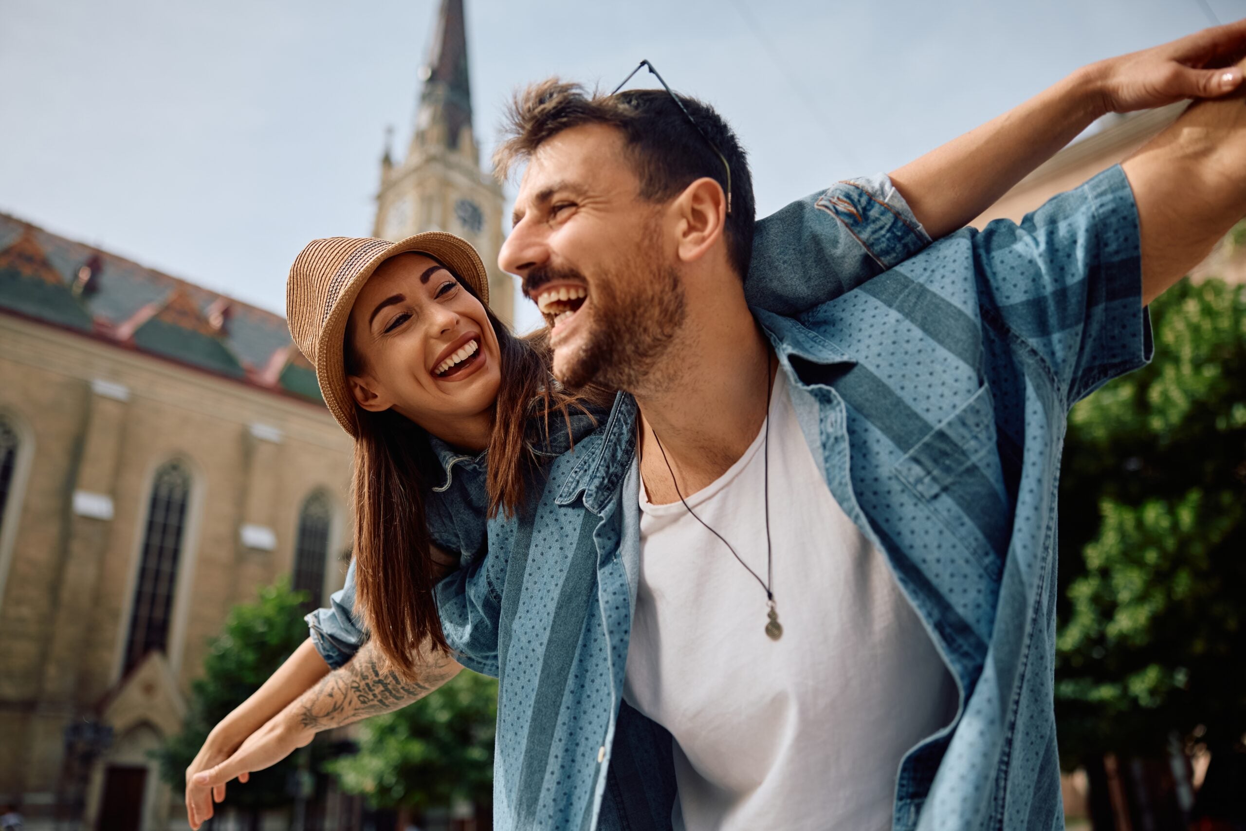 Playful couple of travelers having fun while piggybacking with arms outstretched in the city.