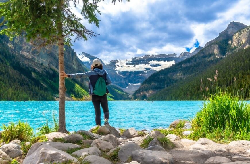 Female tourist standing by a tree on the rocky shore of turquoise lake louise, admiring the stunning view of surrounding mountains and glaciers in banff national park