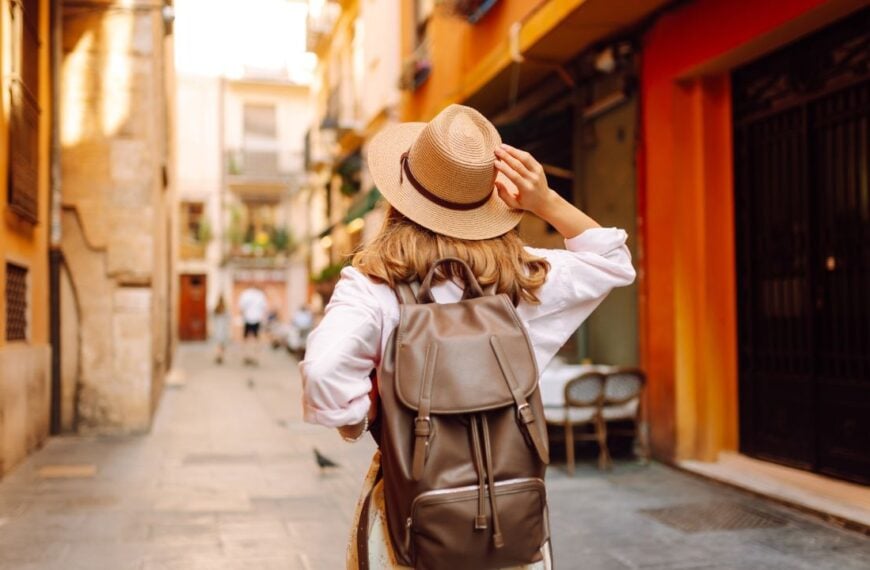 Beautiful tourist wearing hat and carrying backpack strolls through narrow streets of city on sunny day. Young woman enjoys the architecture and the city walk. A weekend, travel concept.