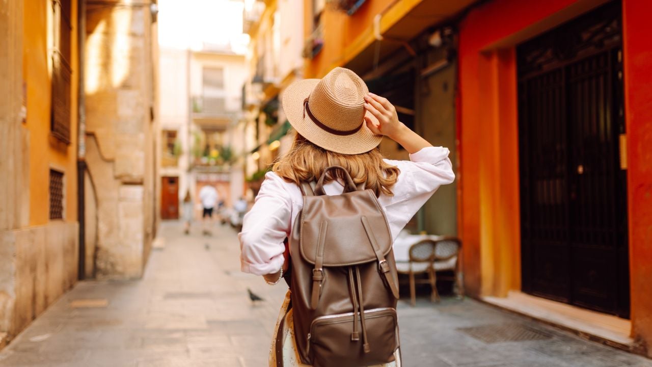 Beautiful tourist wearing hat and carrying backpack strolls through narrow streets of city on sunny day. Young woman enjoys the architecture and the city walk. A weekend, travel concept.