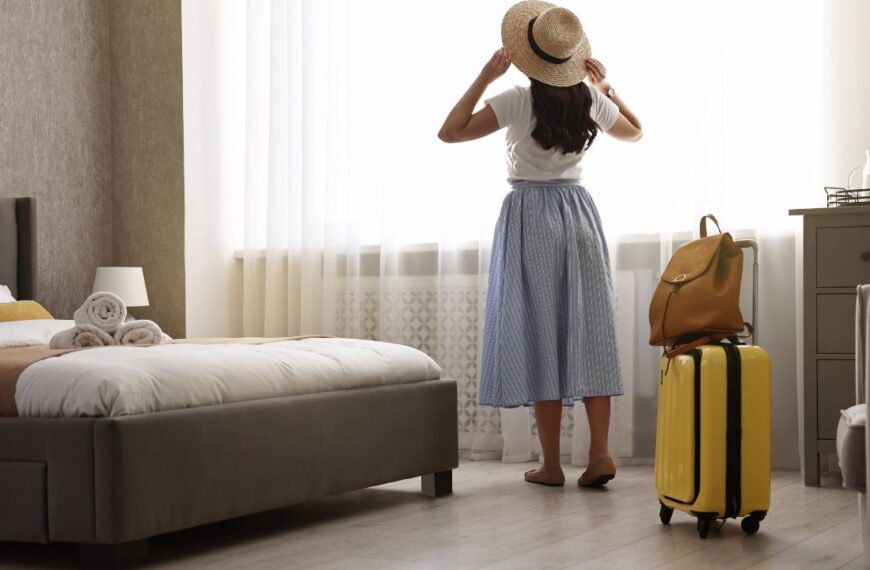 Traveller with suitcase standing near window in hotel room, back view
