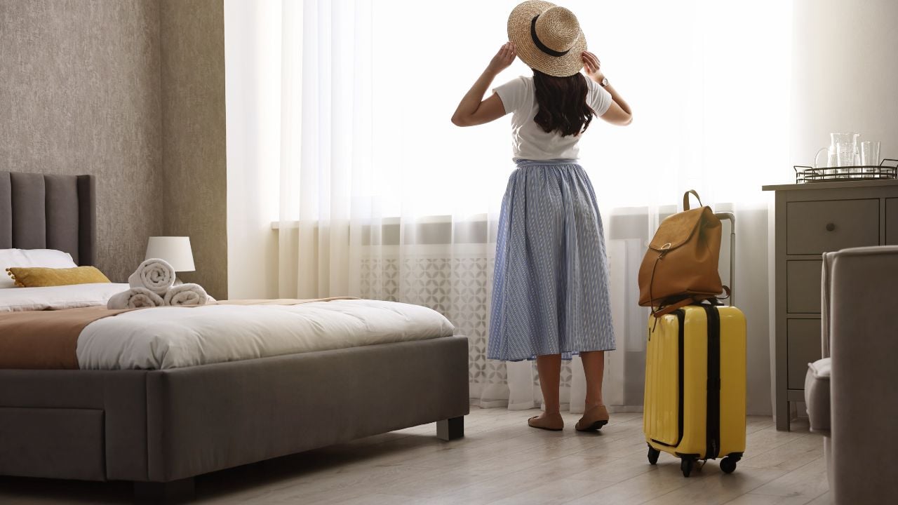 Traveller with suitcase standing near window in hotel room, back view
