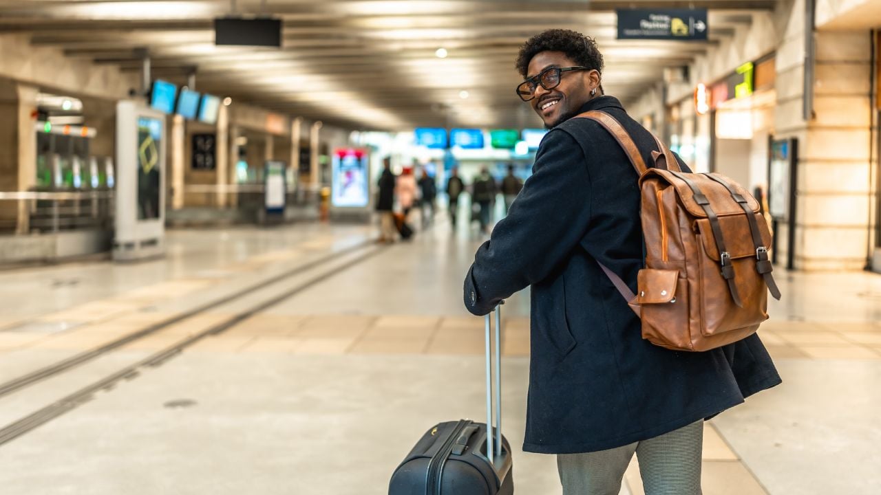 Smiling african man carrying backpack and pulling luggage, looking back while walking through a bright train station, representing business travel and commuting
