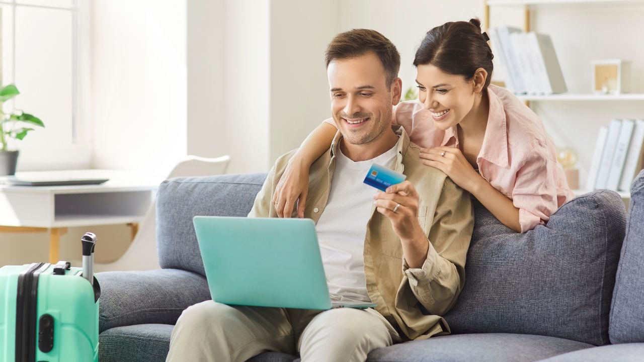 Couple with laptop makes online credit card payment at home. they plan travel, compare deals, and manage costs beside a suitcase. Concept secure payment, holiday trip, vacation and travel booking.