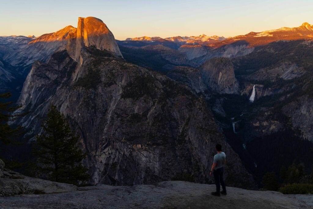 a hiker looks out at half dome at sunset from glacier point